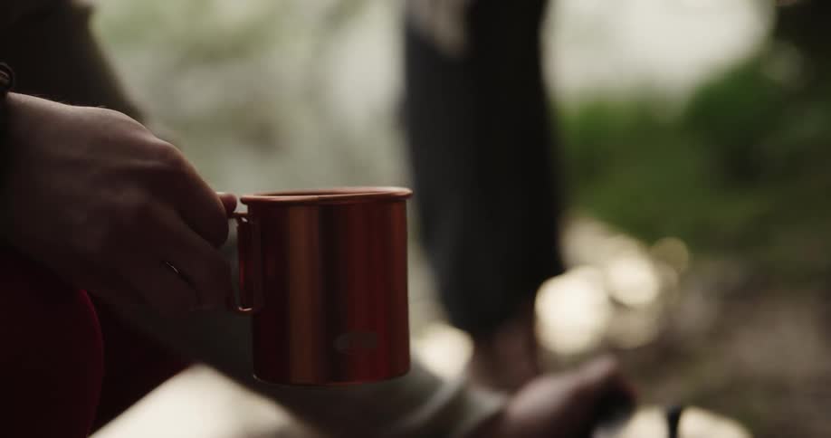 A person pours a liquid drink from a tea pot to a cup beside a river bank