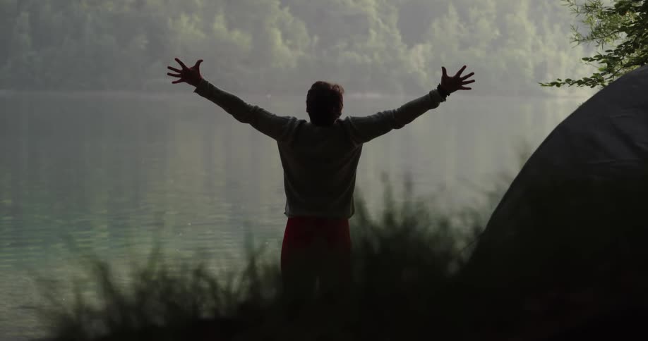 A man walking and stretching by a lake with plants in the foreground and trees in the background
