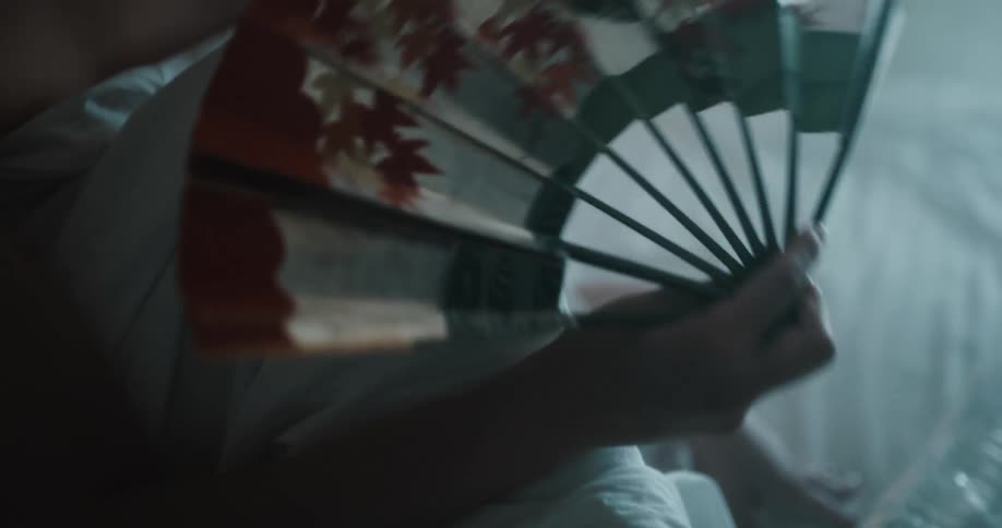 A woman is sitting on her bed and has a fan