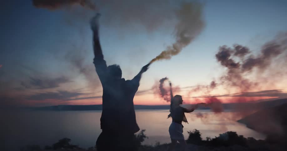 A man and a woman by a lake spinning with cylinders with smoke with the sky and clouds in the background