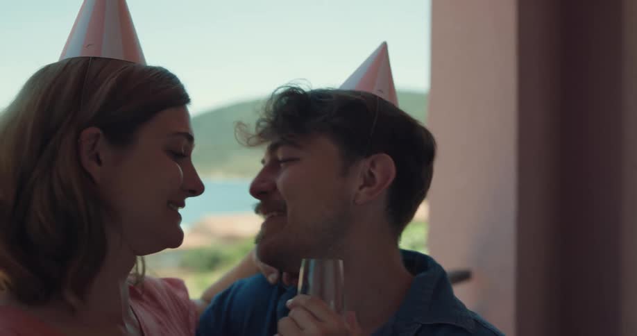 A woman sitting on a mans lap kisses him then rubs her nose against his and puts down a glass of champagne
