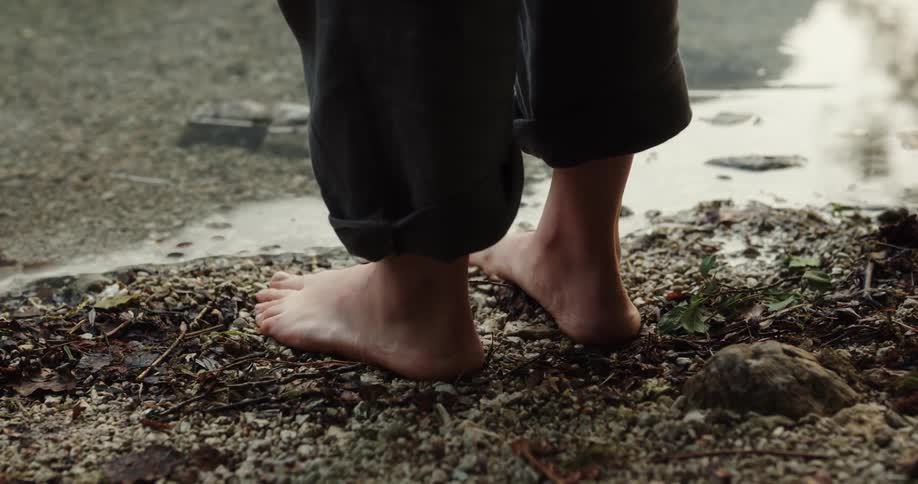 Barefoot woman standing on the shoreline of a lake near a person wearing boots