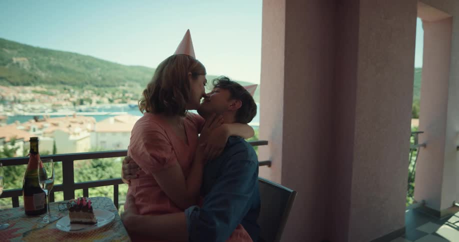A man and a woman sitting on a balcony kissing with a lake the sky and clouds in the background