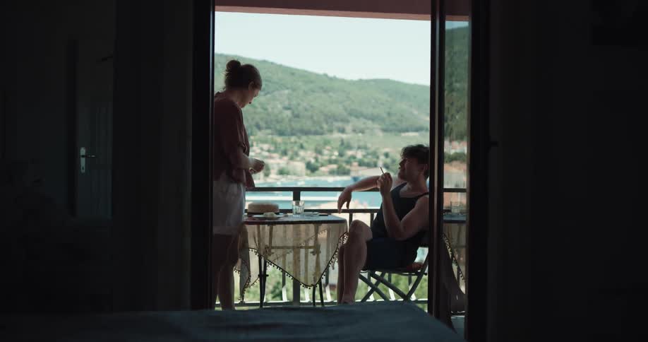 A man lights a cigarette at a table on a balcony and a woman reaches for it