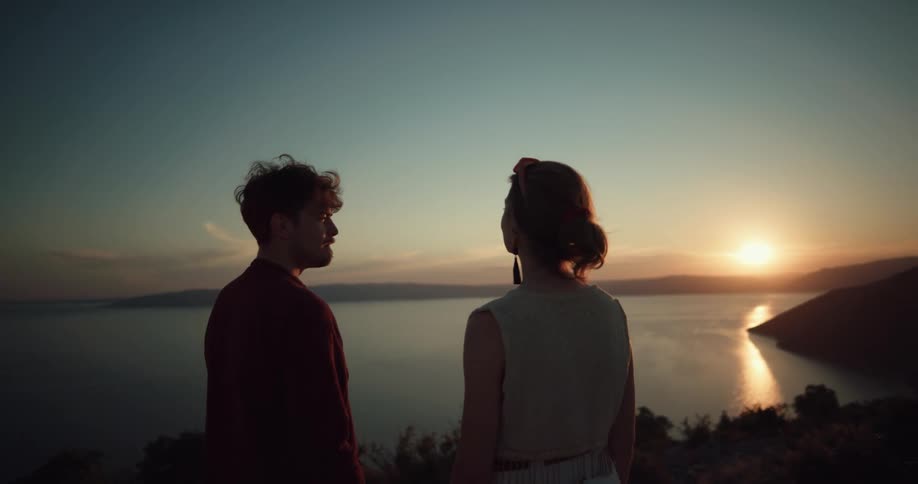 A couple stand on a cliff overlooking the ocean and watch the sun set