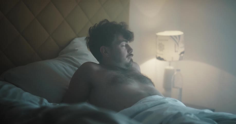 A man wakes up and rubs his eyes as a woman sleeps in bed next to him