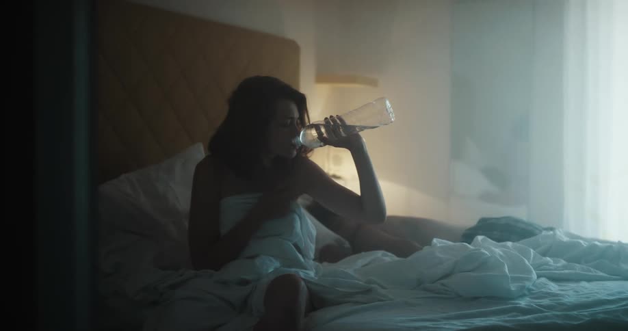 A woman is sitting on a bed drinking from a water bottle while a man lays nearby