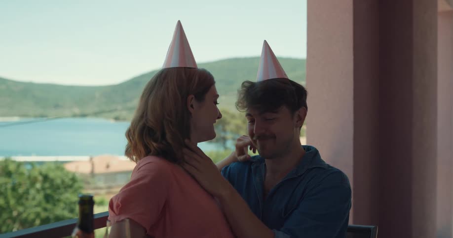 A man and a woman sitting on a balcony drinking and kissing with a lake the sky and clouds in the background