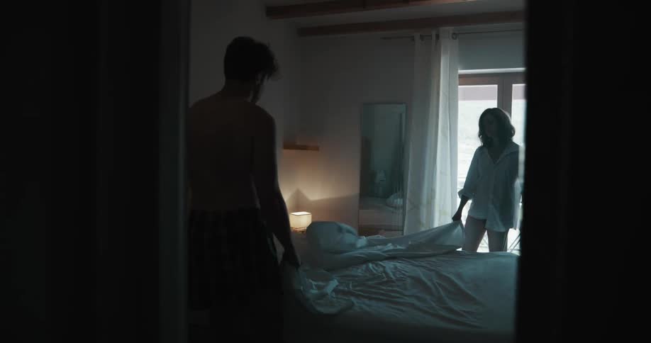 Young couple making the bed inside a dimly lit bedroom in the morning