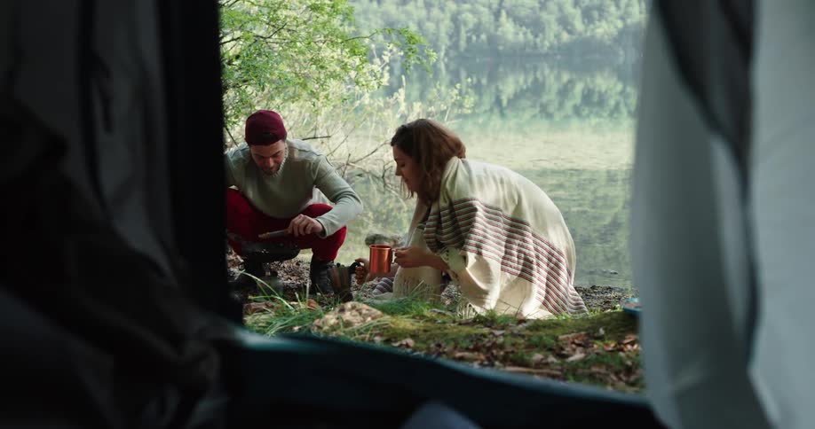 A barefoot female camper kneels beside a man cooking over a campfire as she pours herself a cup of coffee