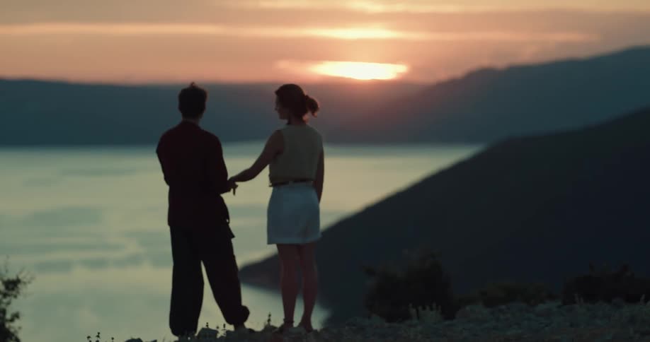 A man and a woman holding hands and standing by a lake with the sky and clouds in the background
