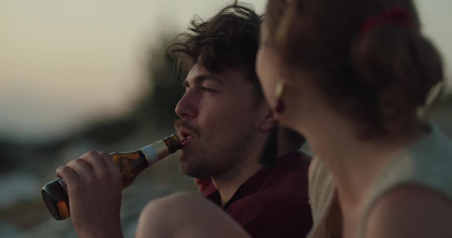 A man and a woman sitting with the man drinking a bottle of beer with the sky and clouds in the background