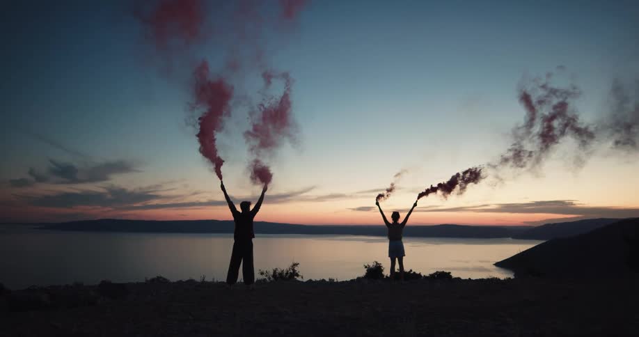 Young couple standing in front of a lake with red smoke flare in their hands at sunset