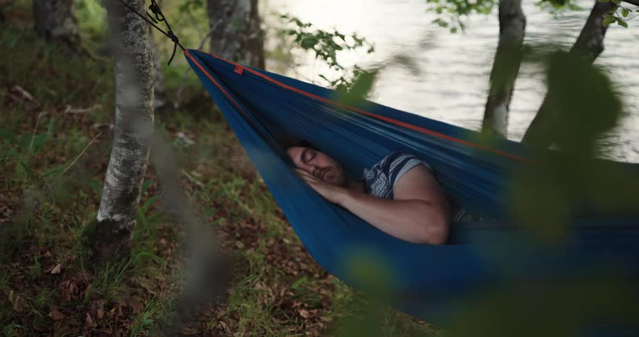Man sleeps in blue hammock next to the lake in the woods