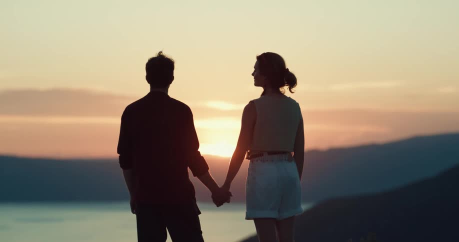 A man and a woman hold hands facing the sunset and a body of water