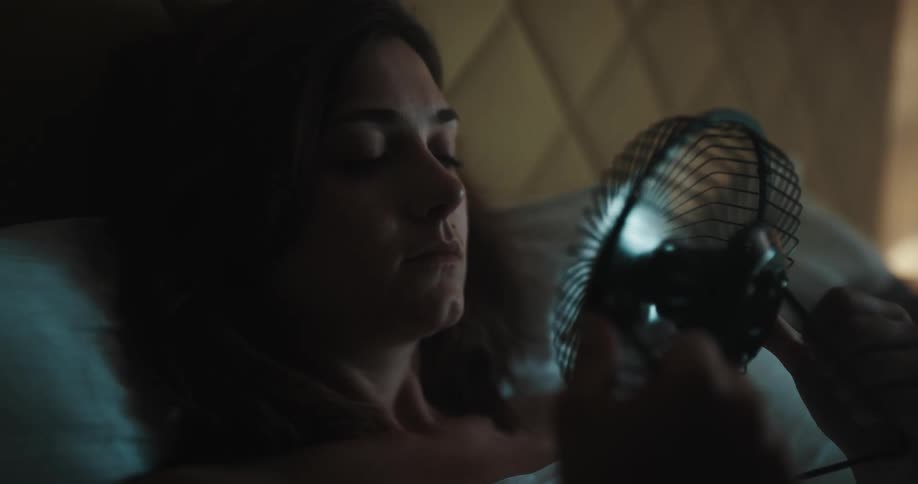 A woman is lying on the bed blowing air on her face with a plugin fan