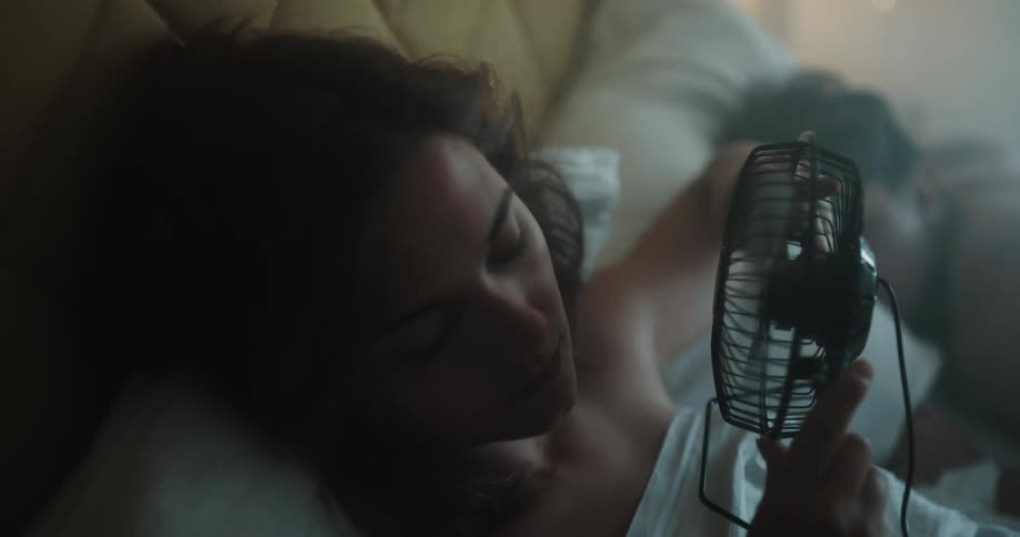 A woman lying in bed next to her boyfriend who is sleeping while she tries to cool off with a mini fan