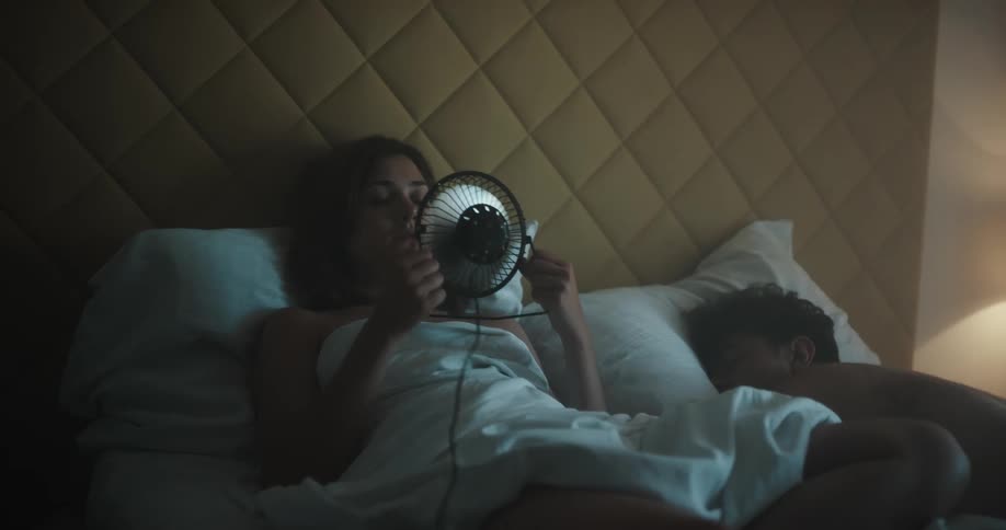 A man and a woman in a bedroom laying in bed with a woman holding a fan