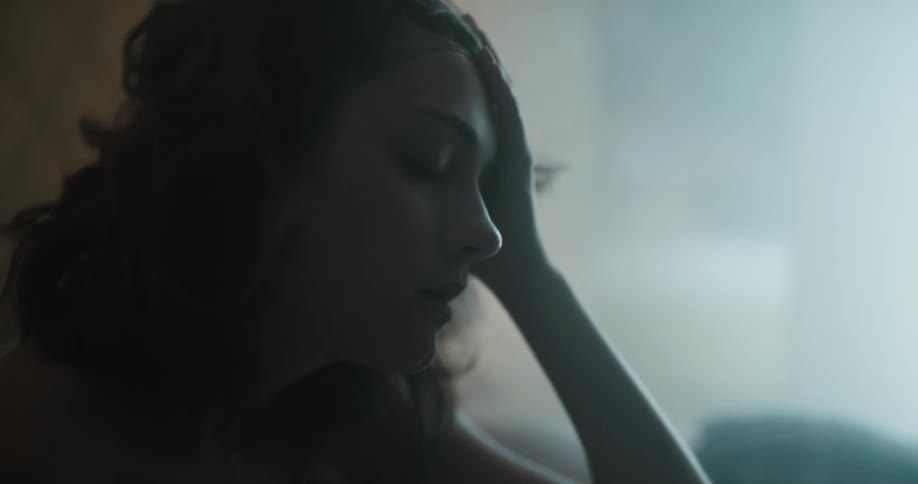 A woman sitting up in bed with sheet wrapped around her rubs her face and head as if in pain