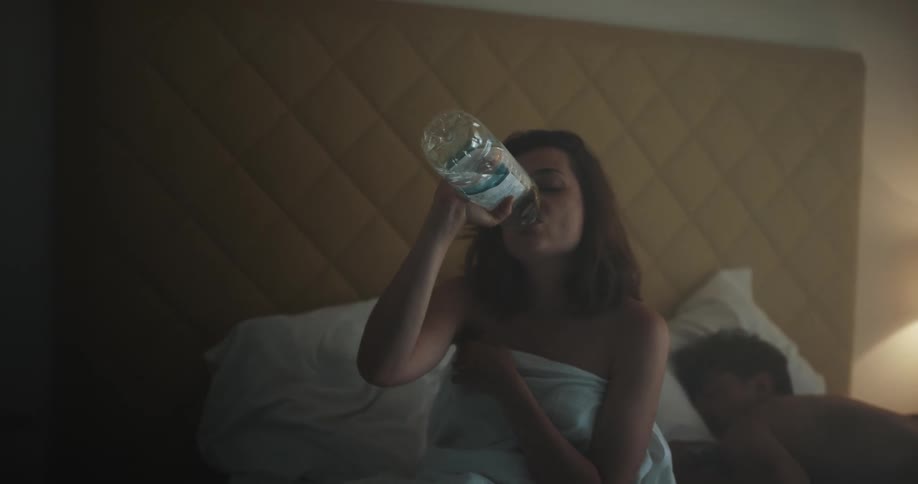 A woman takes a drink of water from a bottle then gets up from a bed a man is sleeping in