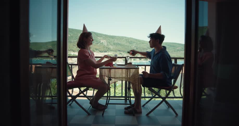 A man and a woman are sitting on a balcony pouring champagne