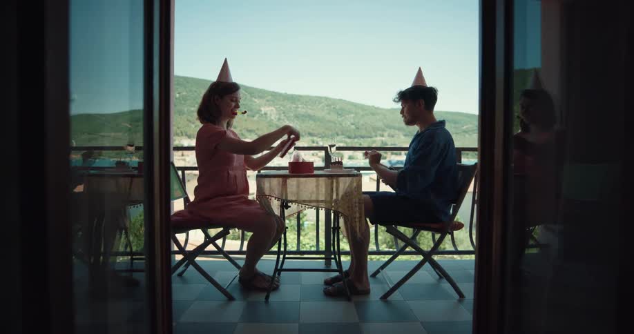 A man and woman sit at a table on a balcony wearing party hats and opening a bottle of champagne