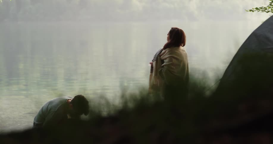 Woman stands stretching on shore of lake near tent as man leans near ground