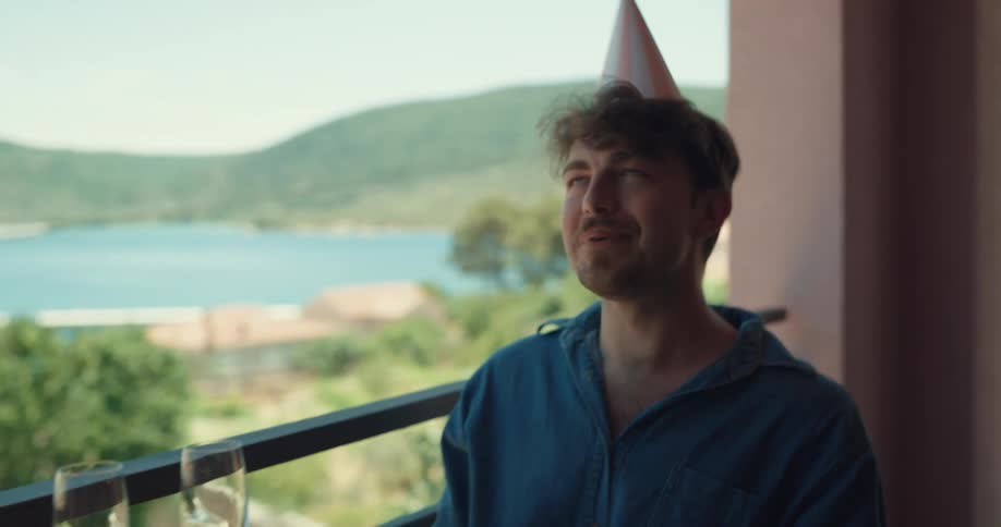 A man with a party hat on a balcony sitting at a table and feeding a woman with a lake the sky and clouds in the background