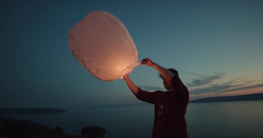 Man lighting a paper balloon in front of a lake at sunset