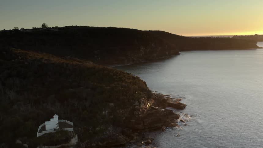A forest and buildings sit on the side of the mountain as the ocean waves roll into shore at dawn