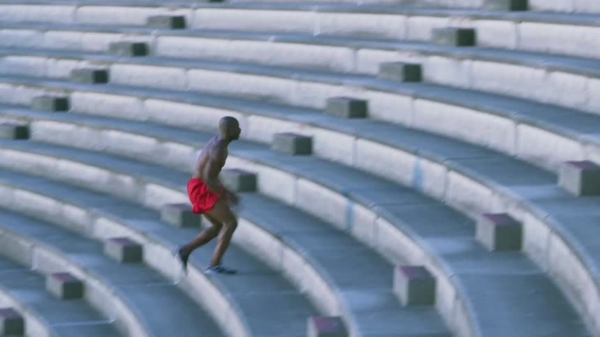 A man, wearing only shorts, enthusiastically runs up concrete bleachers in a stadium