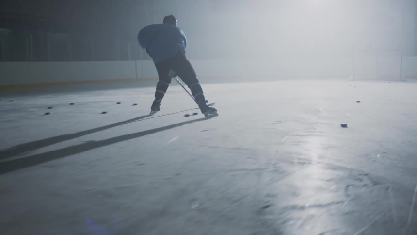 A hockey player practices shots in dark ice rink lit by a bright light