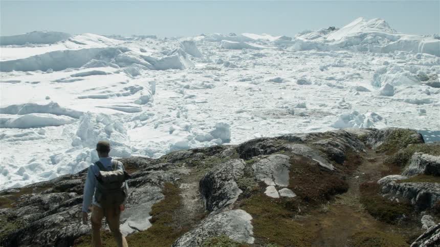 A man wearing a backpack walks over rocky landscape towards snow