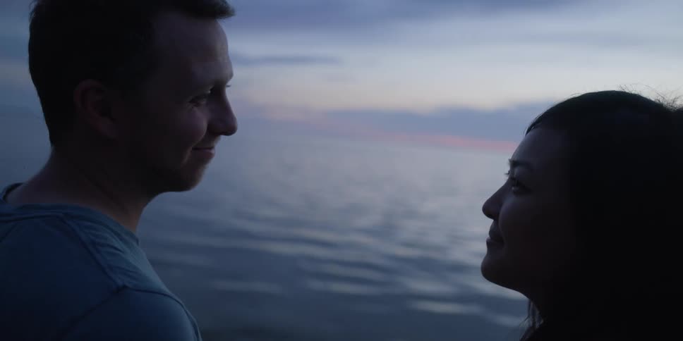 A man and woman smile and look into one anothers eyes while standing on a beach at sunset