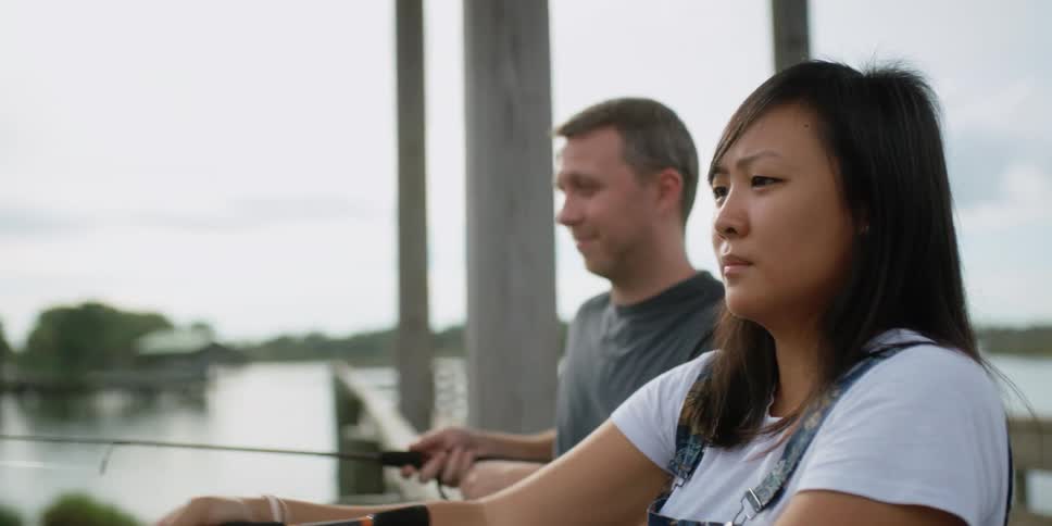 A woman casts her fishing line into a lake while standing on a wooden pier next to a man who is also fishing