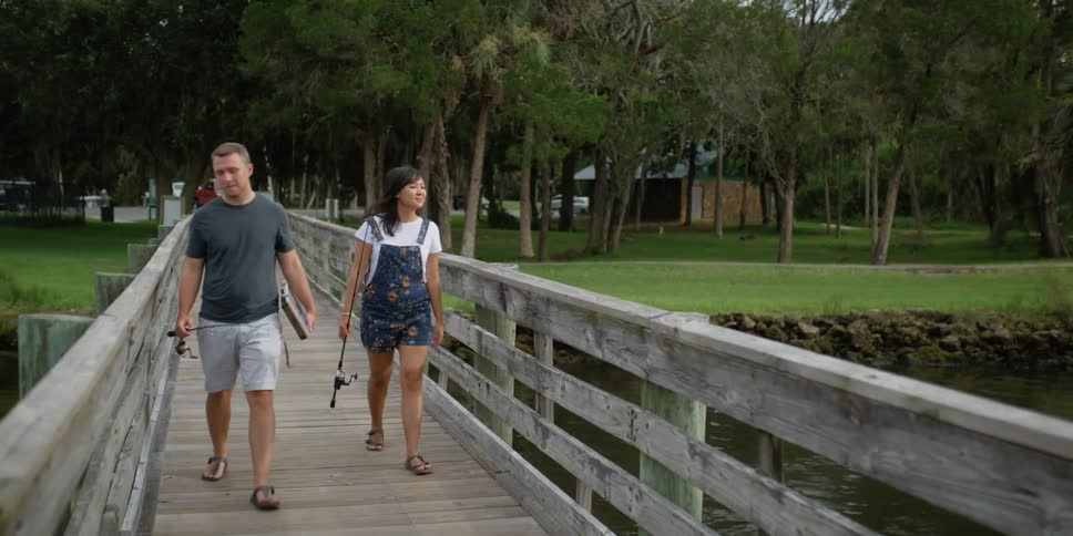 A man and woman walk along a wooden dock while carrying fishing rods