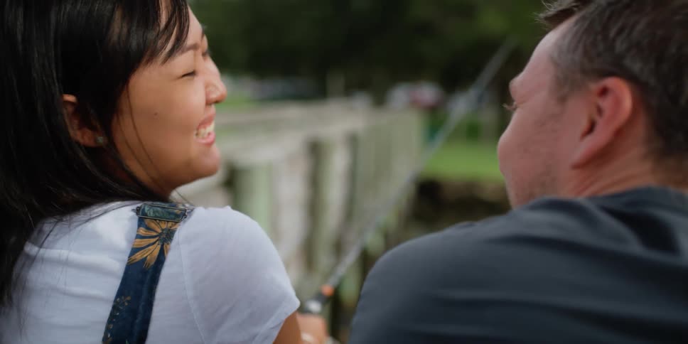 A man and woman laugh and chat with fishing together on the side of a river during the day