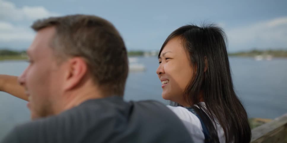 A woman standing on a pier on a lake points out something in the distance to a man fishing next to her