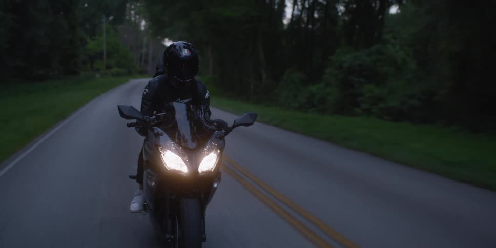 Cyclist wearing a black jacket and black helmet is riding a motorcycle with the headlights on down a road surrounded by trees