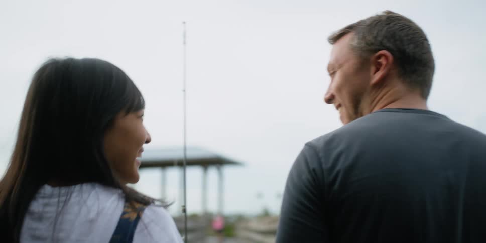 A man and woman walk together towards a dock while the woman carries a fishing rod