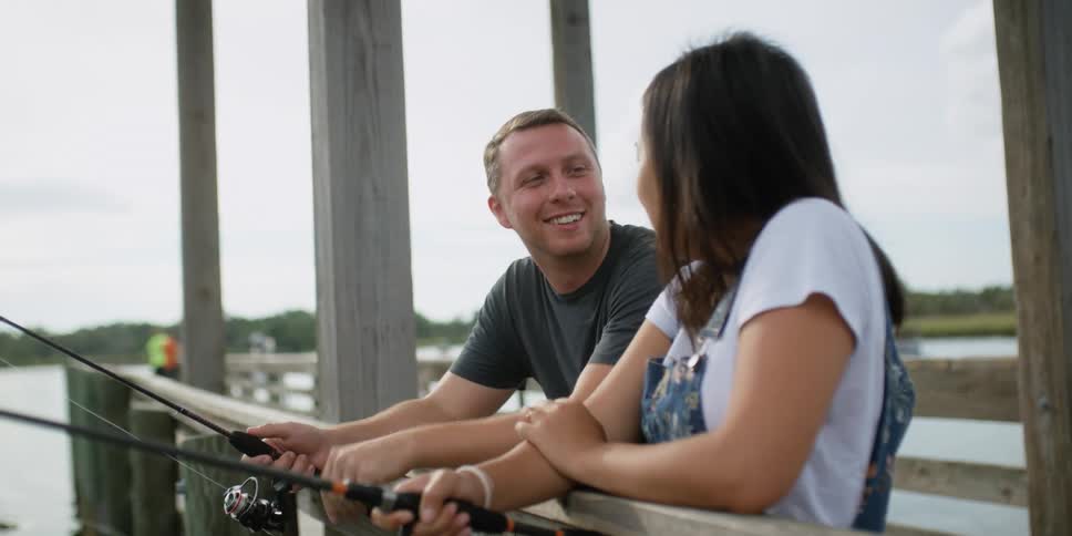 A man touches his girlfriends face as they stand on a pier and fish at a lake