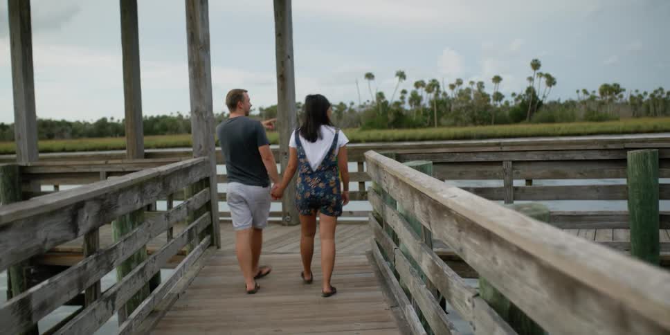 A couple walks along a pier while holding hands and pointing out something in the distance