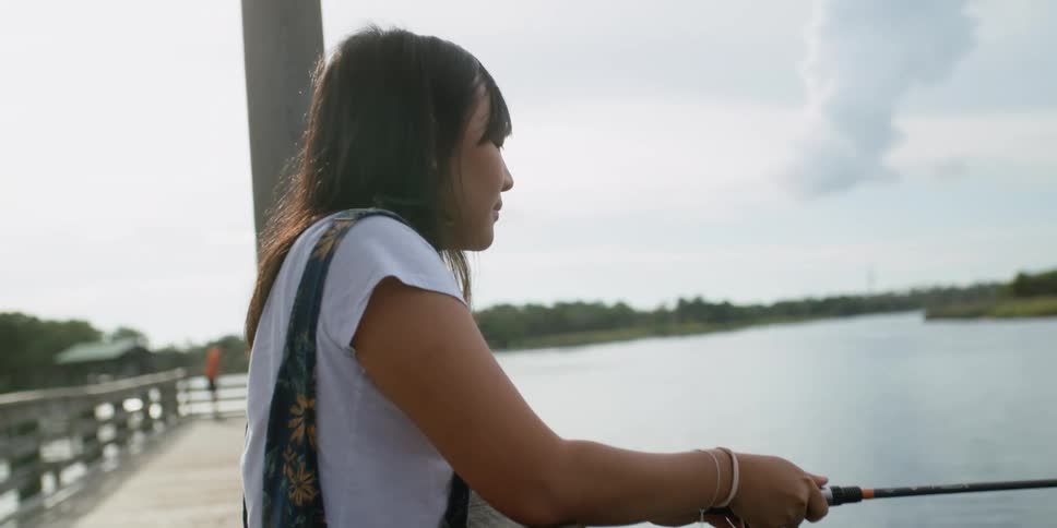 A woman on a dock by a lake fishing with a fishing pole with trees the sky and clouds in the background