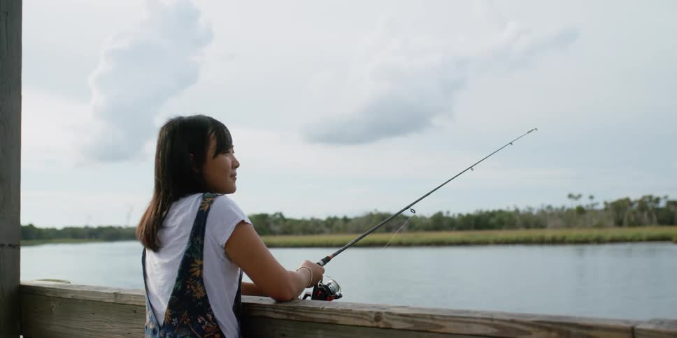 A woman stands on a pier holding a fishing pole to the lake