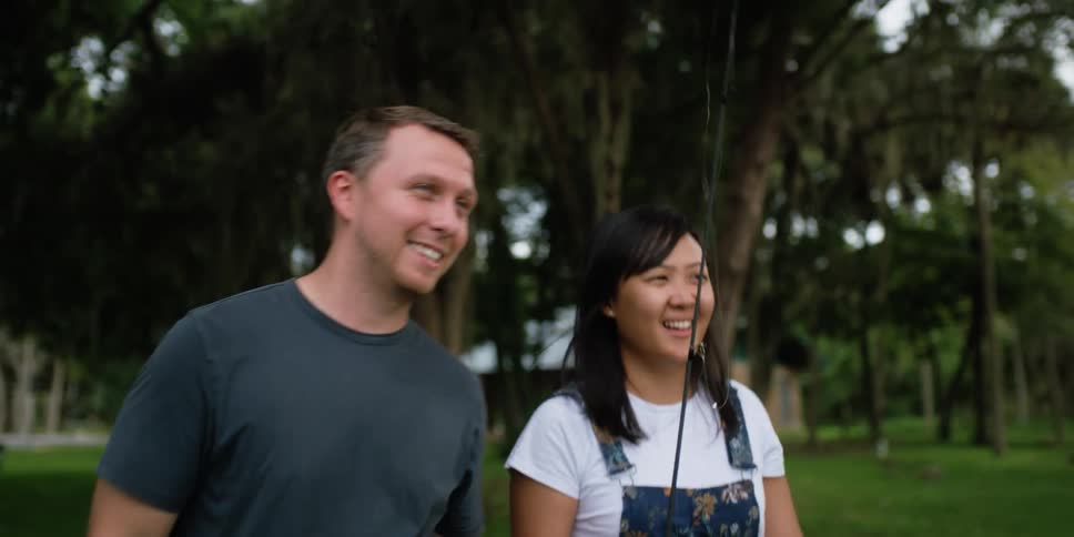 A man and woman walk together through a grass park while the woman carries a fishing rod