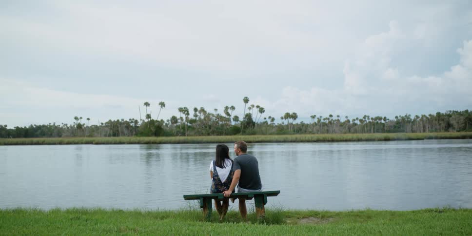 A man and woman chat while sitting together on a bench on the shore of a lake during the day