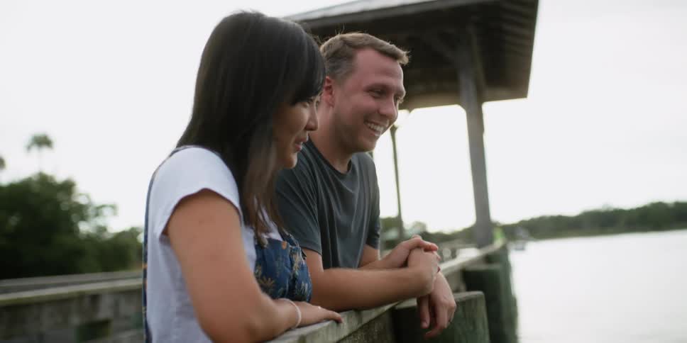 A man and woman hold hands while leaning against a dock railing talking and smiling