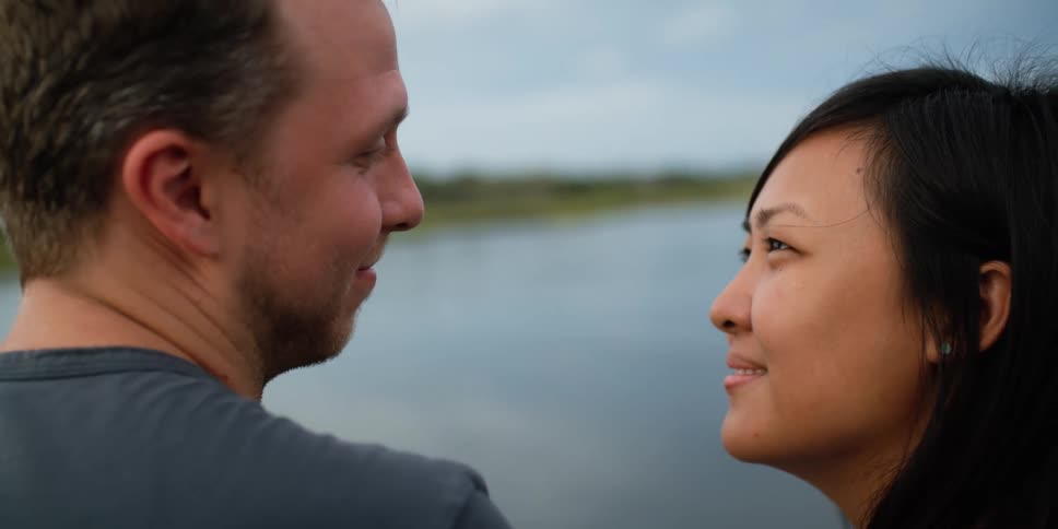 A man and woman standing by a lake with trees the sky and clouds in the background