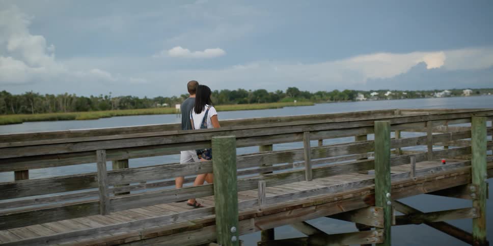 A man and woman hold hands and walk to the end of a pier on a lake