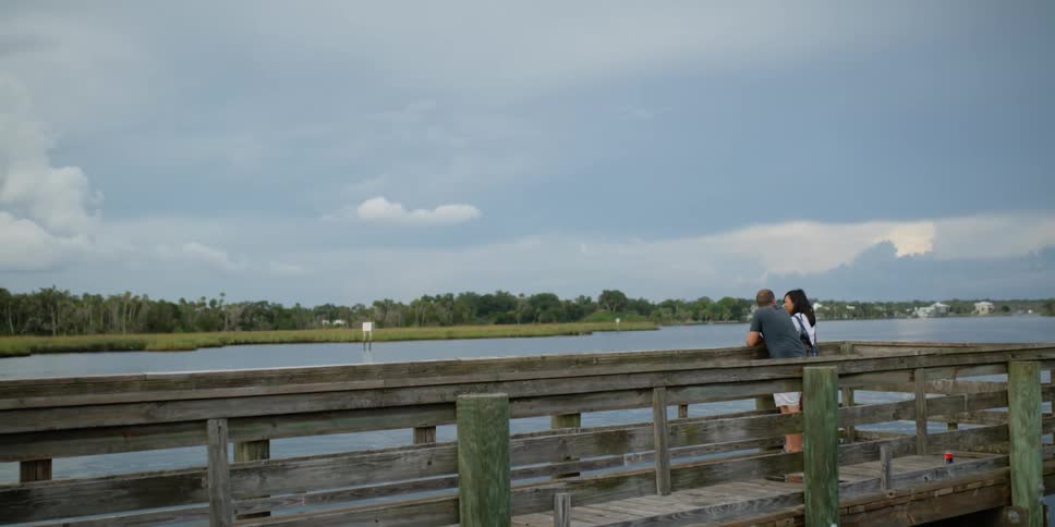 A man and woman are standing and talking on a wooden bridge next to a lake